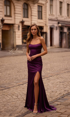 A woman wearing the Strapless Long Satin Prom Dress: La Femme 32952 with a thigh-high slit stands on a cobblestone street in front of historic buildings.