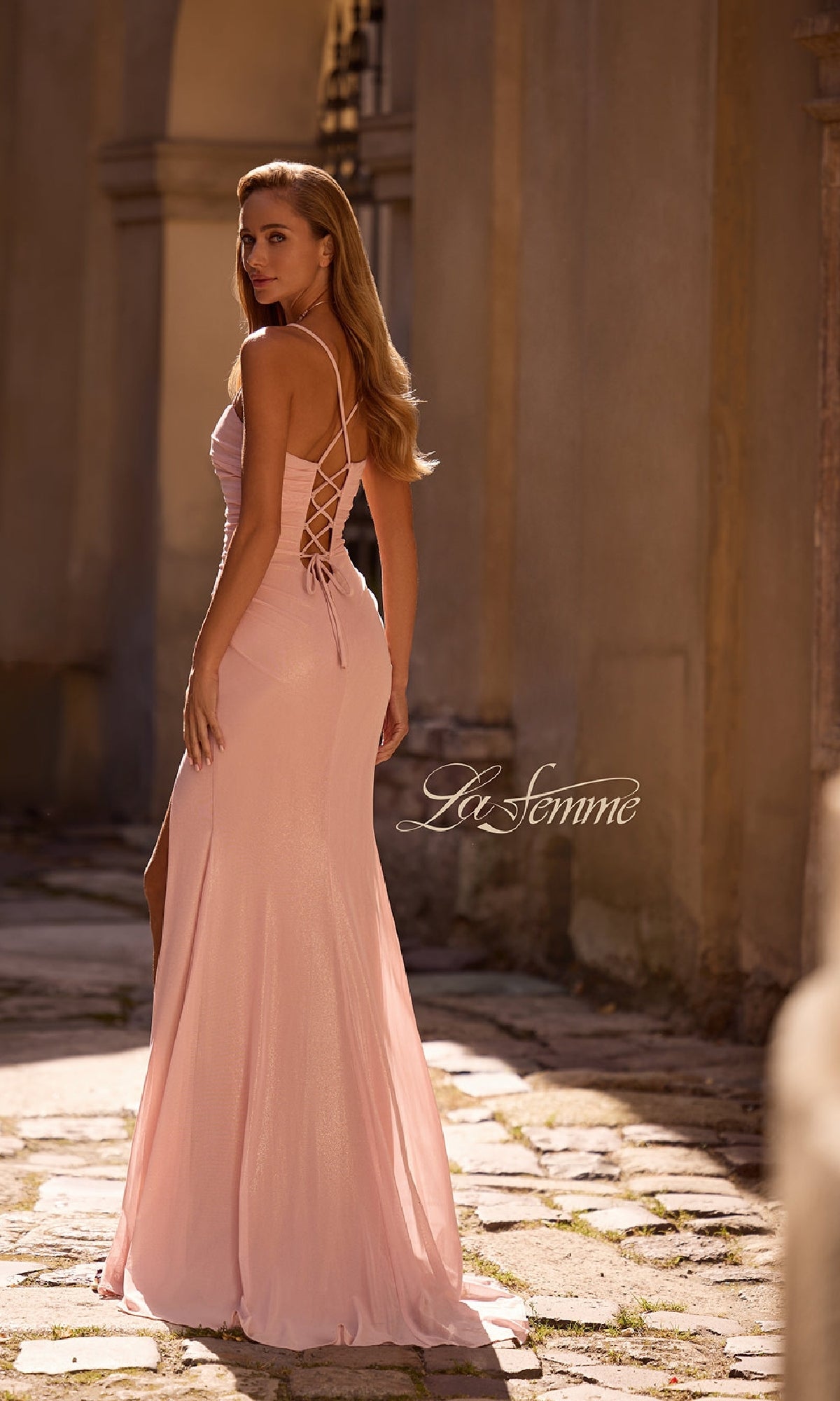 Woman wearing the La Femme 32964 long prom dress in light pink with strappy lace-up back and subtle sweetheart neckline, standing outdoors on a stone path and looking over her shoulder. The 