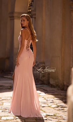 Woman wearing the La Femme 32964 long prom dress in light pink with strappy lace-up back and subtle sweetheart neckline, standing outdoors on a stone path and looking over her shoulder. The "La Femme" logo is visible.