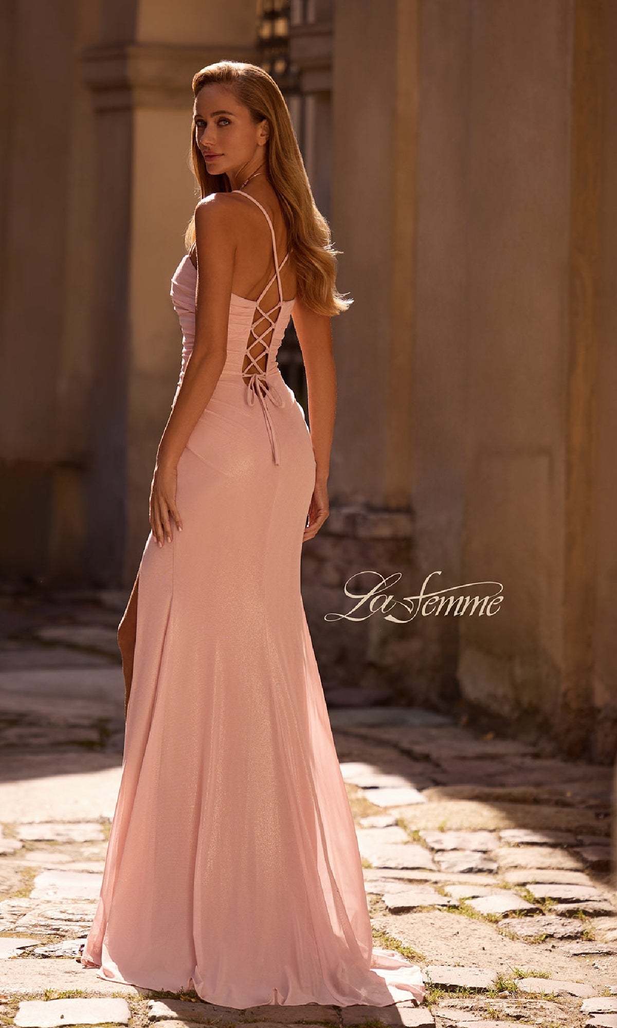A woman wears the La Femme 32964 long prom dress—a light pink, sleeveless gown with a sweetheart neckline and lace-up back—while standing outdoors on a stone pathway, glancing over her shoulder. The 