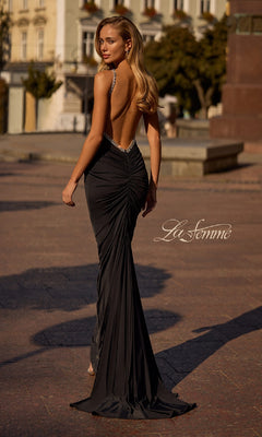 A woman stands outdoors, looking over her shoulder in a black Backless Ruched Long Prom Dress: La Femme 32970. The long gown features jeweled straps, a ruched design, slight train, and the visible “La Femme” logo.
