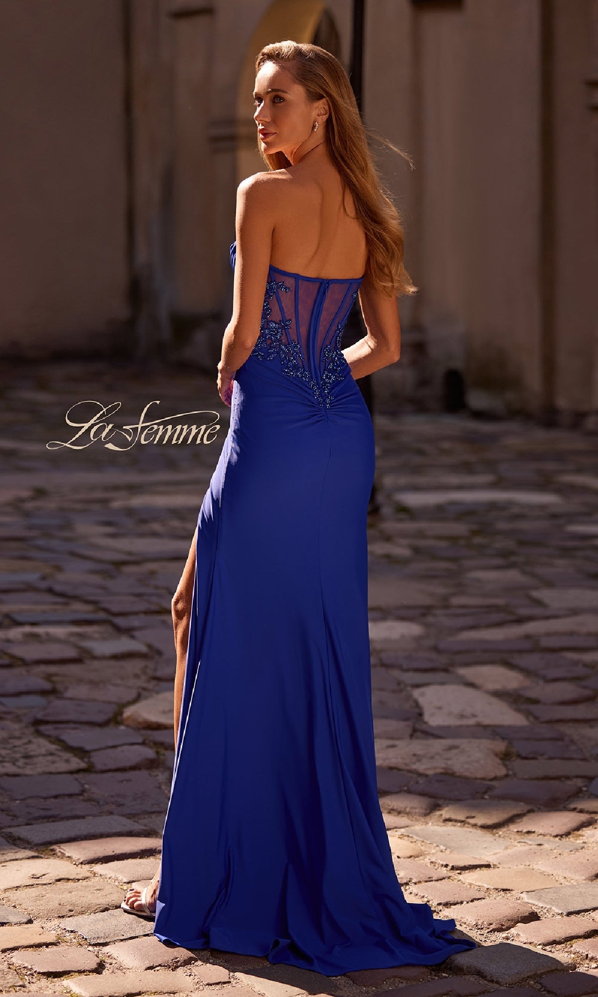 A woman stands on a cobblestone street facing away, wearing the Beaded-Bodice Long Prom Dress: La Femme 32981 in royal blue, featuring a strapless neckline, sheer lace back, beaded designs, and a high slit.