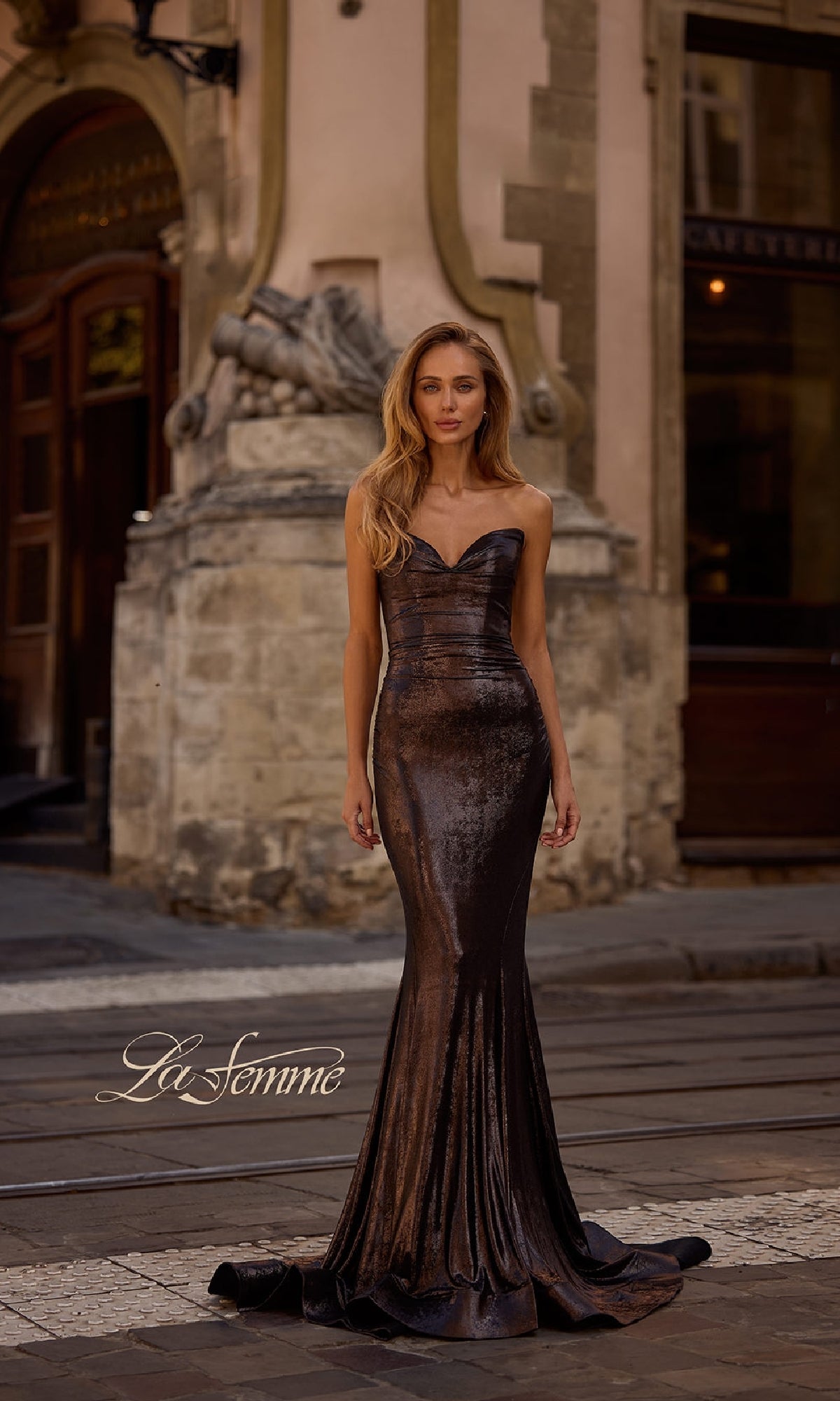 A woman in the La Femme 32995 strapless long metallic prom dress stands on a cobblestone street before an ornate stone building, with the 