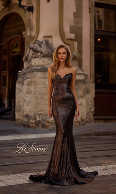 A woman in the La Femme 32995 strapless long metallic prom dress stands on a cobblestone street before an ornate stone building, with the "La Femme" logo in the lower left corner.