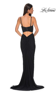 A woman stands facing away, wearing La Femme 32996—a long black prom dress with thin straps, a low back, ruched detailing, a small train, and cut-out accents for a timelessly elegant silhouette.