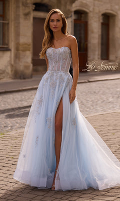 A woman stands gracefully on a cobblestone street wearing the La Femme 33028, a strapless beaded long prom gown in light blue with a corset bodice, rhinestone floral appliques, and a high slit.