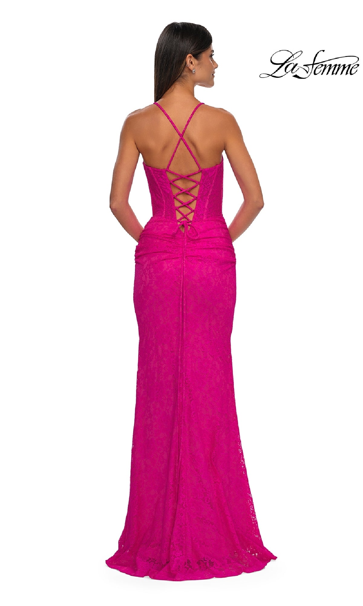 Woman in a bright pink Long Stretch-Lace Prom Dress: La Femme 33036, featuring crisscross straps, a lace-up back, and a corset-bodice design, stands facing away from the camera.