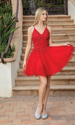 A woman in the Lace-Up Short Babydoll Homecoming Dress 3303, featuring a red, sleeveless design with sequin details and a corset bodice, stands on a brick path holding out one side of her skirt.