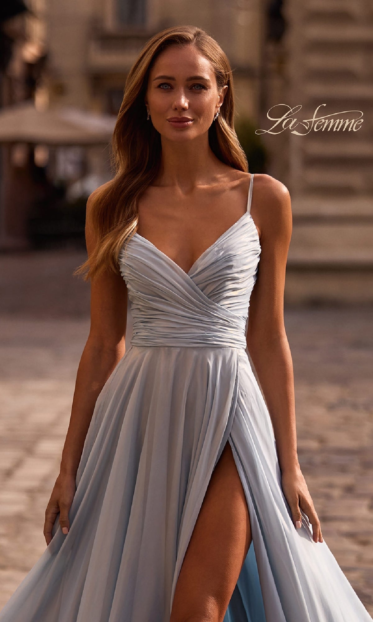 A woman walks outdoors on a cobblestone street in the La Femme 33043 Metallic Chiffon Long Prom Dress, featuring a light blue hue, thigh-high slit, and pleated bodice for added elegance.