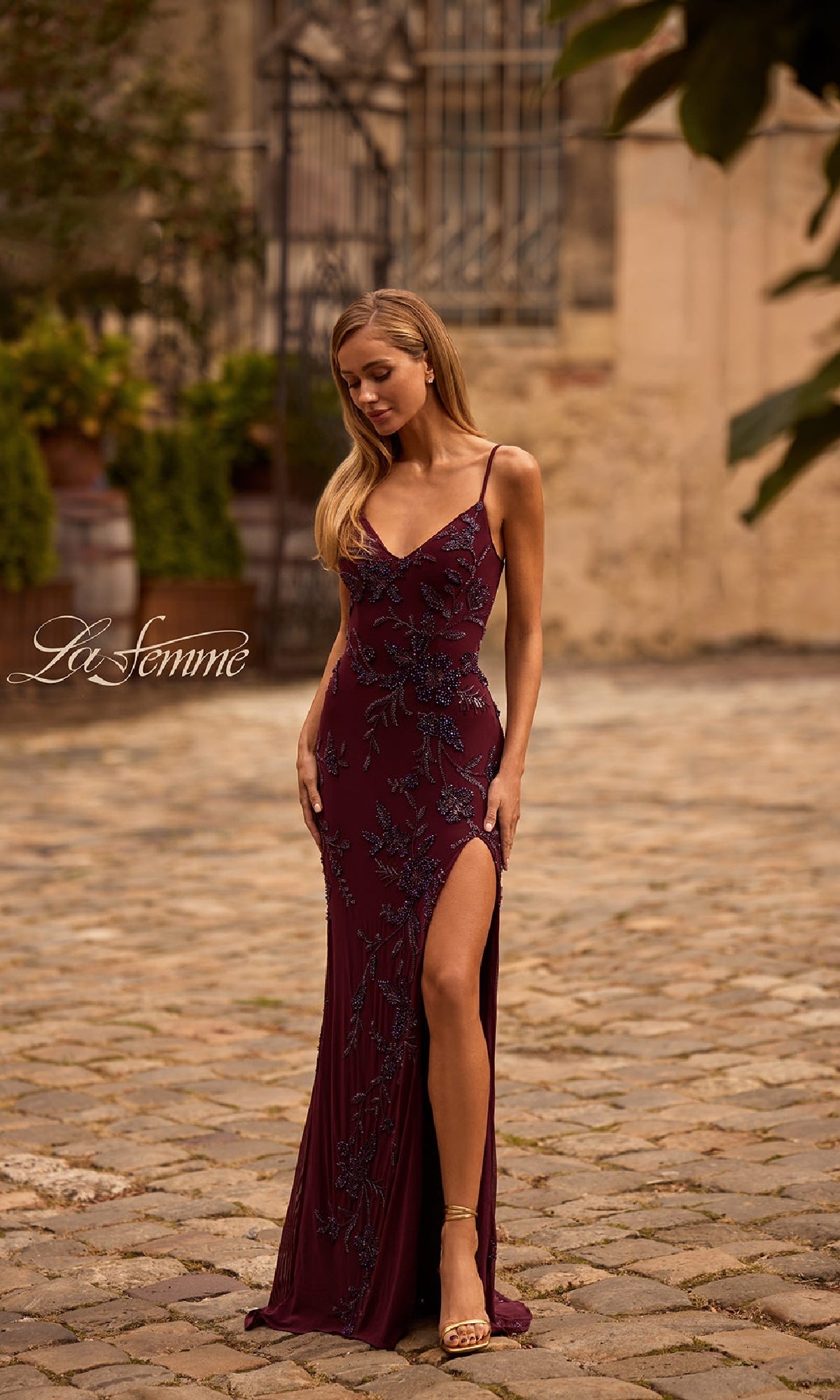 A woman stands outdoors on a cobblestone path wearing the Lace-Up Long Beaded Prom Dress: La Femme 33054—a burgundy evening gown with a thigh-high slit, floral embellishments, and thin shoulder straps for an elegant look.