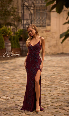 A woman stands outdoors on a cobblestone path wearing the Lace-Up Long Beaded Prom Dress: La Femme 33054—a burgundy evening gown with a thigh-high slit, floral embellishments, and thin shoulder straps for an elegant look.