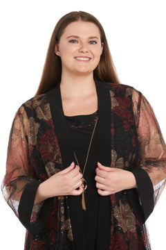 A woman smiles, wearing the R&M Richards 3312W Plus-Size Jacket Party Dress with a sheer floral-patterned jacket over a black top, holding the lapels; white background.