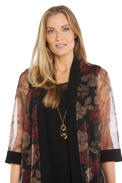A woman with long blonde hair models the R&M Richards 3312 Petite Two-Piece Party Dress, featuring a black semi-formal style and sheer floral-patterned shawl, accessorized with a long pendant necklace against a white background.