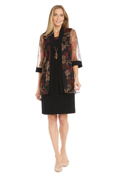 Woman wearing the R&M Richards 3312 Petite Two-Piece Party Dress, featuring a black knee-length semi-formal dress with a sheer floral-patterned jacket, and nude heels, standing and smiling at the camera.