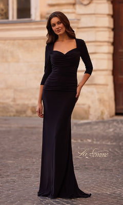 A woman stands outdoors on cobblestone wearing the La Femme 33135 long black jersey formal dress with three-quarter sleeves and a sweetheart neckline, exuding timeless elegance.