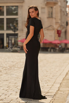 A woman stands on a cobblestone street, looking over her shoulder in the La Femme 33146 long formal dress with elegant ruching. Buildings and red umbrellas are visible in the background.