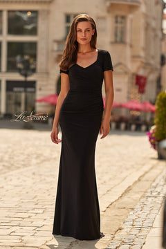 A woman in a fitted, short-sleeved black La Femme 33146 long formal dress with elegant ruching stands on a cobblestone street in an urban setting during the day.