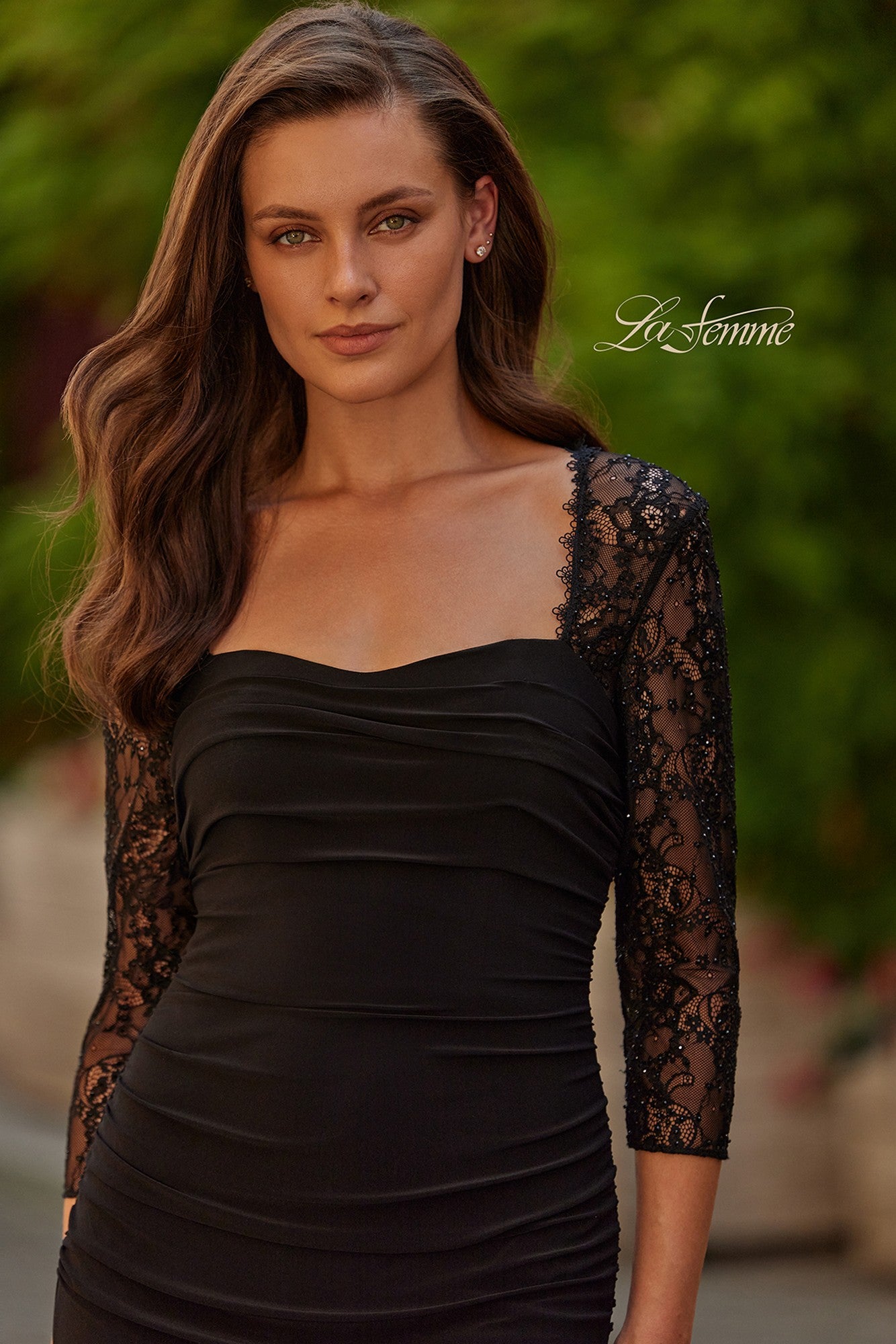 A woman with long brown hair wears the La Femme 33147 long formal dress, a fitted black gown with lace illusion sleeves, while standing outdoors in front of greenery.