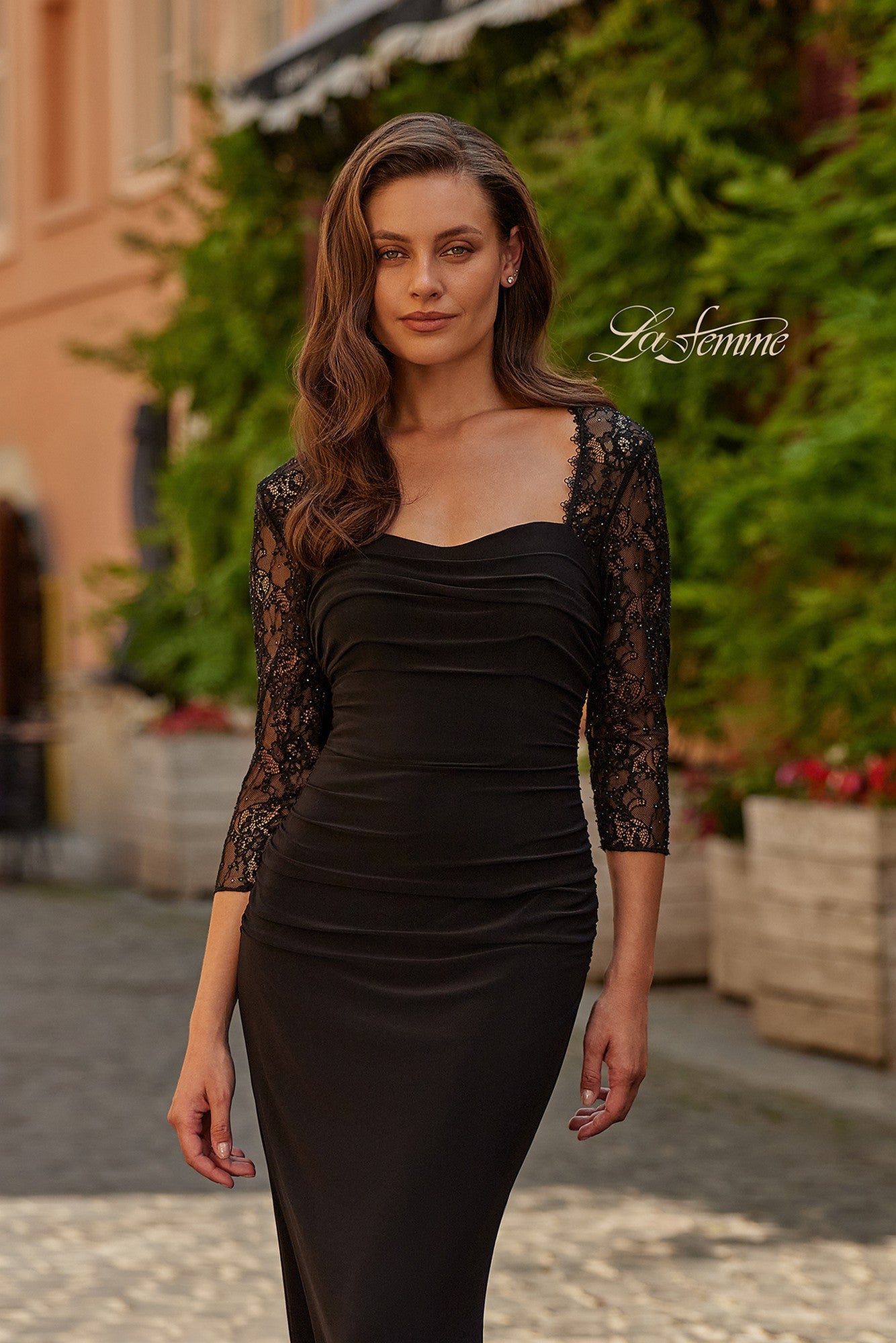 Wearing the La Femme 33147 long formal dress with fitted black silhouette and lace illusion sleeves, a woman stands on a cobblestone street with greenery and buildings behind her—her elegant look perfect for a formal evening.