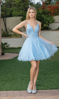 A woman stands on grass in a light blue Short Babydoll Homecoming Dress with beading and silver high heels, with greenery and a wall in the background.