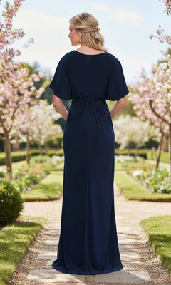 A woman with braided hair stands on a garden path, seen from behind in the La Femme 33158 long prom dress—navy blue, floor-length with short sleeves and a beaded waist belt.