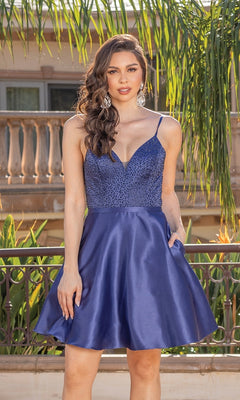 A woman poses outside in a Short A-Line Homecoming Dress With Lace-Up Back, featuring a deep blue satin finish, beaded v-neck bodice, and flared skirt, set against greenery and a balcony railing.