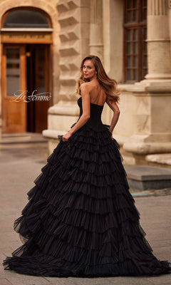 A woman wearing the La Femme 33202 long black strapless prom dress with a pleated bodice and ruffled skirt stands outdoors before an ornate building, glancing over her shoulder.