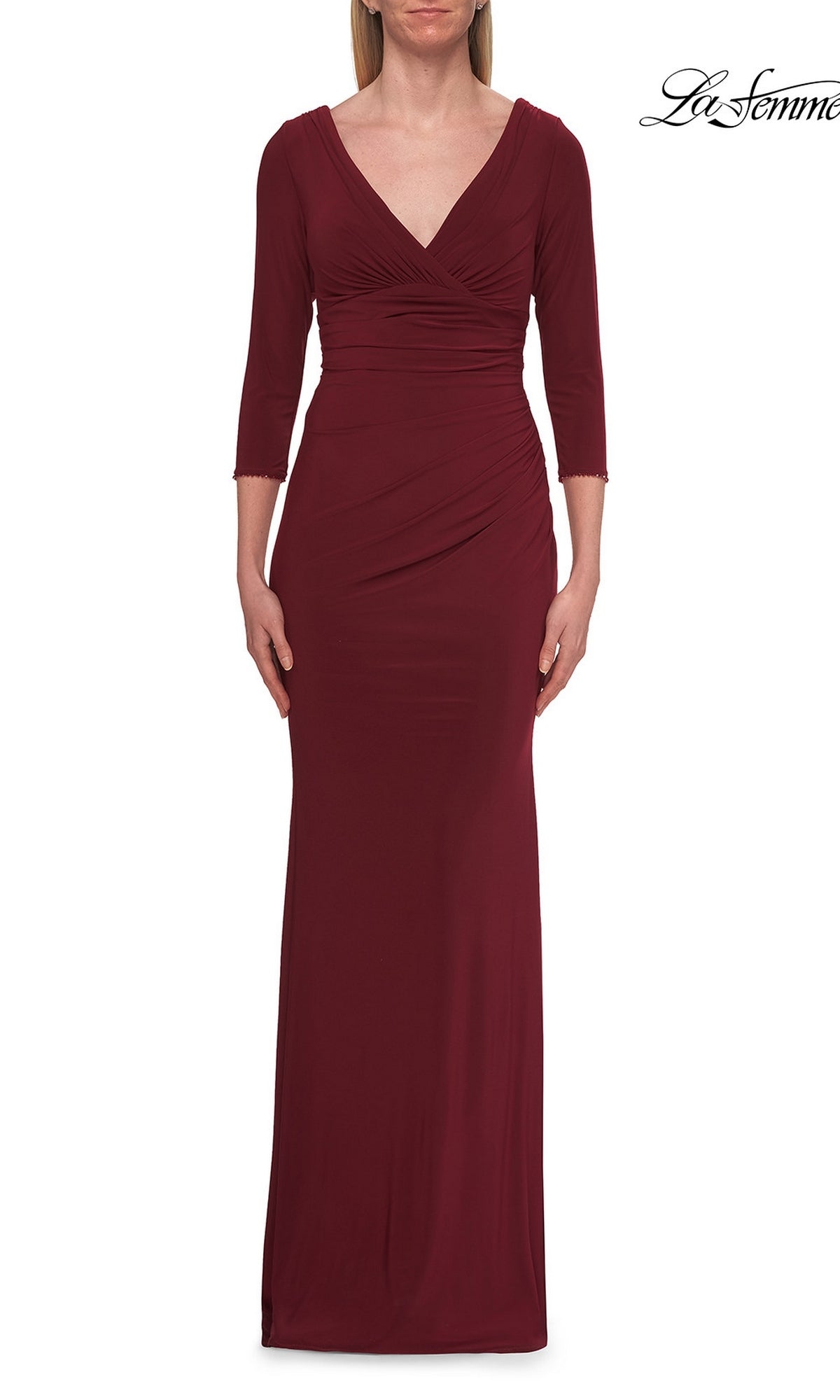 A person wearing the La Femme 33214 Long Sleeve Jersey Formal Gown in wine, featuring three-quarter sleeves, a V-neckline, and gathered waist detailing, stands against a white background.