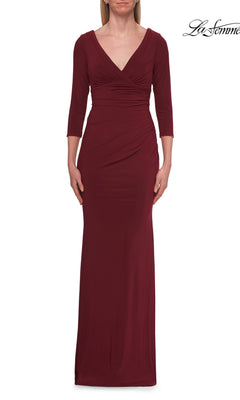 A person wearing the La Femme 33214 Long Sleeve Jersey Formal Gown in wine, featuring three-quarter sleeves, a V-neckline, and gathered waist detailing, stands against a white background.