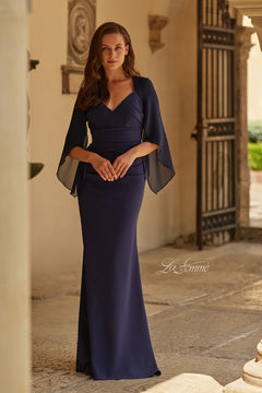 A woman stands in an outdoor corridor wearing the La Femme 33215, a floor-length navy formal dress with a beaded V-neckline and chiffon flutter sleeves.