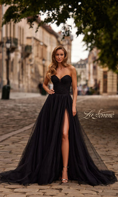 A woman with long wavy hair and heels stands on a cobblestone street in a black strapless high-slit gown, the La Femme 33307 Long Prom Dress. The "La Femme" logo is visible.