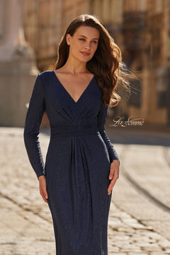 A woman wearing the La Femme 33310, a long-sleeved navy blue formal dress with a ruched waist, stands outdoors on a sunlit cobblestone street.