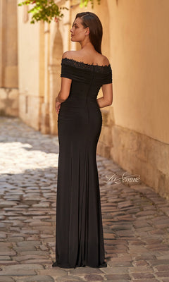 A woman in the Long Prom Dress: La Femme 33329 with a figure-hugging silhouette and off-the-shoulder neckline stands on a cobblestone path, facing away from the camera.