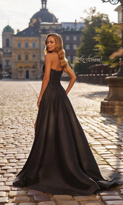 A woman wearing the La Femme 33353 long black Mikado strapless prom dress stands on a cobblestone street, glancing over her shoulder, with historic buildings and a clear sky in the background.