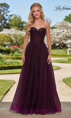 A woman poses elegantly outdoors in the La Femme 33363 long prom dress, a dark purple strapless gown with a fitted corset bodice and flowing tulle skirt, surrounded by greenery and blooming trees.