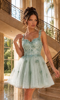 A woman stands indoors wearing the Embroidered-Lace Short Babydoll Hoco Dress 3338 in sage, featuring an embroidered-lace bodice and tulle skirt. Ornate staircase and large windows appear in the background.