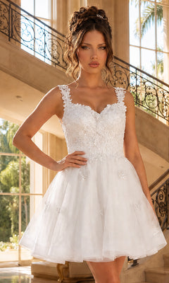 A woman in the Off White Embroidered-Lace Short Babydoll Hoco Dress 3338, featuring a floral appliqué bodice, stands indoors by a staircase and large windows.