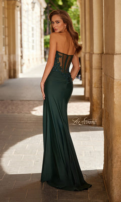 Wearing the La Femme 33396, a floor-length, dark green strapless prom dress with a sheer embroidered back, a woman stands outdoors by stone columns, glancing over her shoulder.