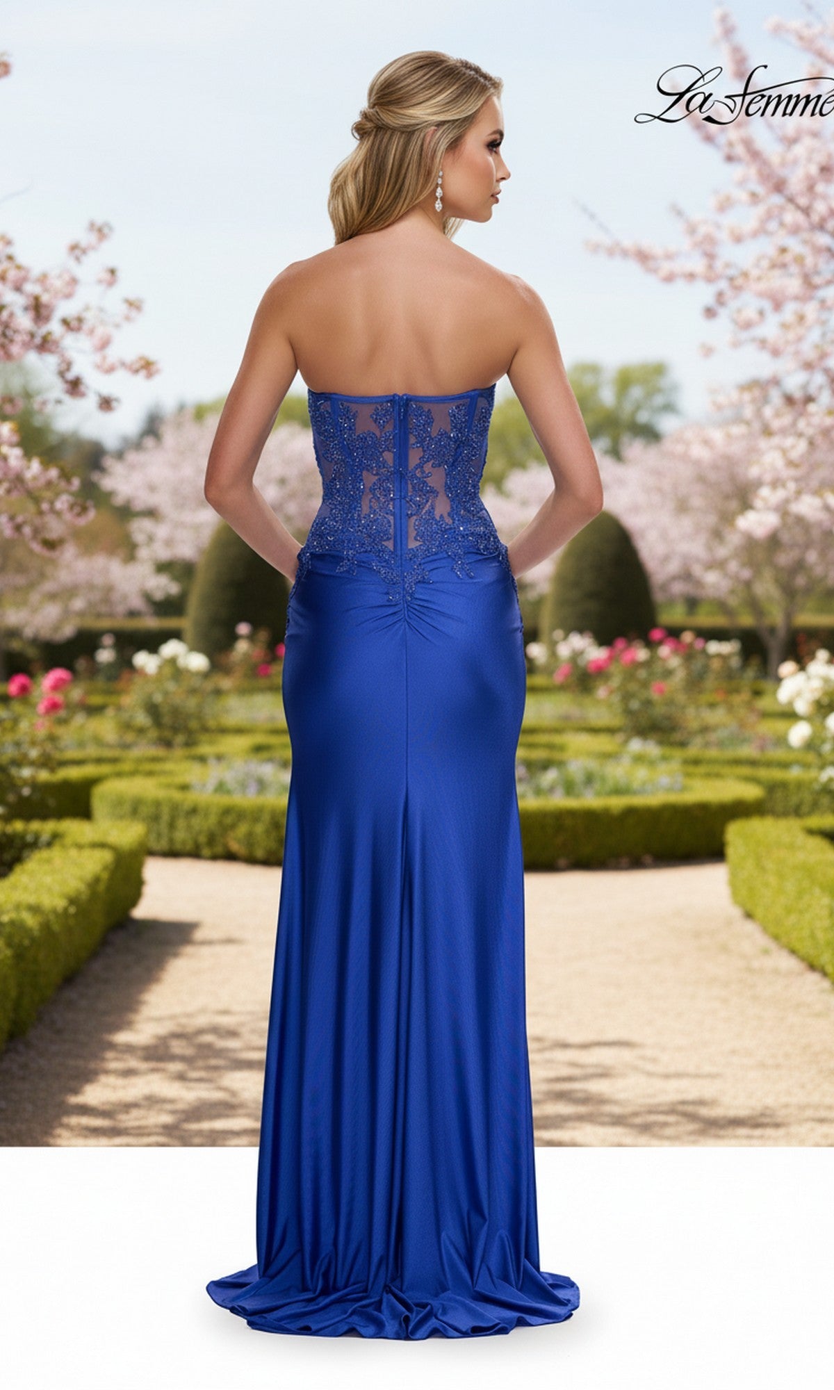 A woman stands outdoors in a manicured garden, wearing the La Femme 33398 long prom dress: a strapless, floor-length royal blue gown with a lace corset bodice detailing the back.