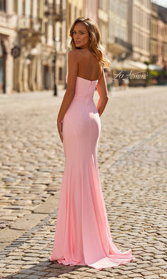 A woman stands on a cobblestone street wearing the Classic Strapless Long Prom Dress, La Femme 33403, in light pink and looks over her shoulder.