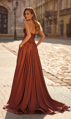 A woman wearing the La Femme 33417 long prom dress, designed with rust-colored satin and a strapless neckline, stands on a cobblestone street, glancing over her shoulder.