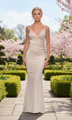 A woman in a fitted Champagne Long Satin Prom Dress with Train, La Femme 33421, stands on a garden path lined with hedges and blooming trees under a clear sky.