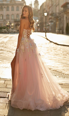 A woman in the La Femme 33432 long prom dress, a strapless light pink ball gown with delicate floral appliqué, stands on a sunlit cobblestone street and looks over her shoulder.