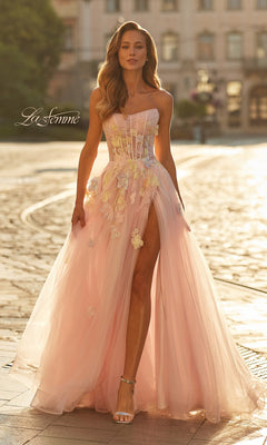 A woman stands on a sunlit cobblestone street wearing the La Femme 33432 long prom dress in light pink, featuring a strapless neckline, high slit, and floral appliqué.