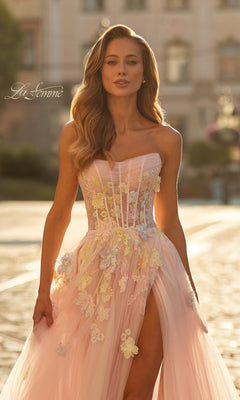 Woman in the La Femme 33432 long prom dress—strapless, pastel pink with floral appliqué and a thigh-high slit—standing outdoors in sunlight; "La Femme" logo in corner.