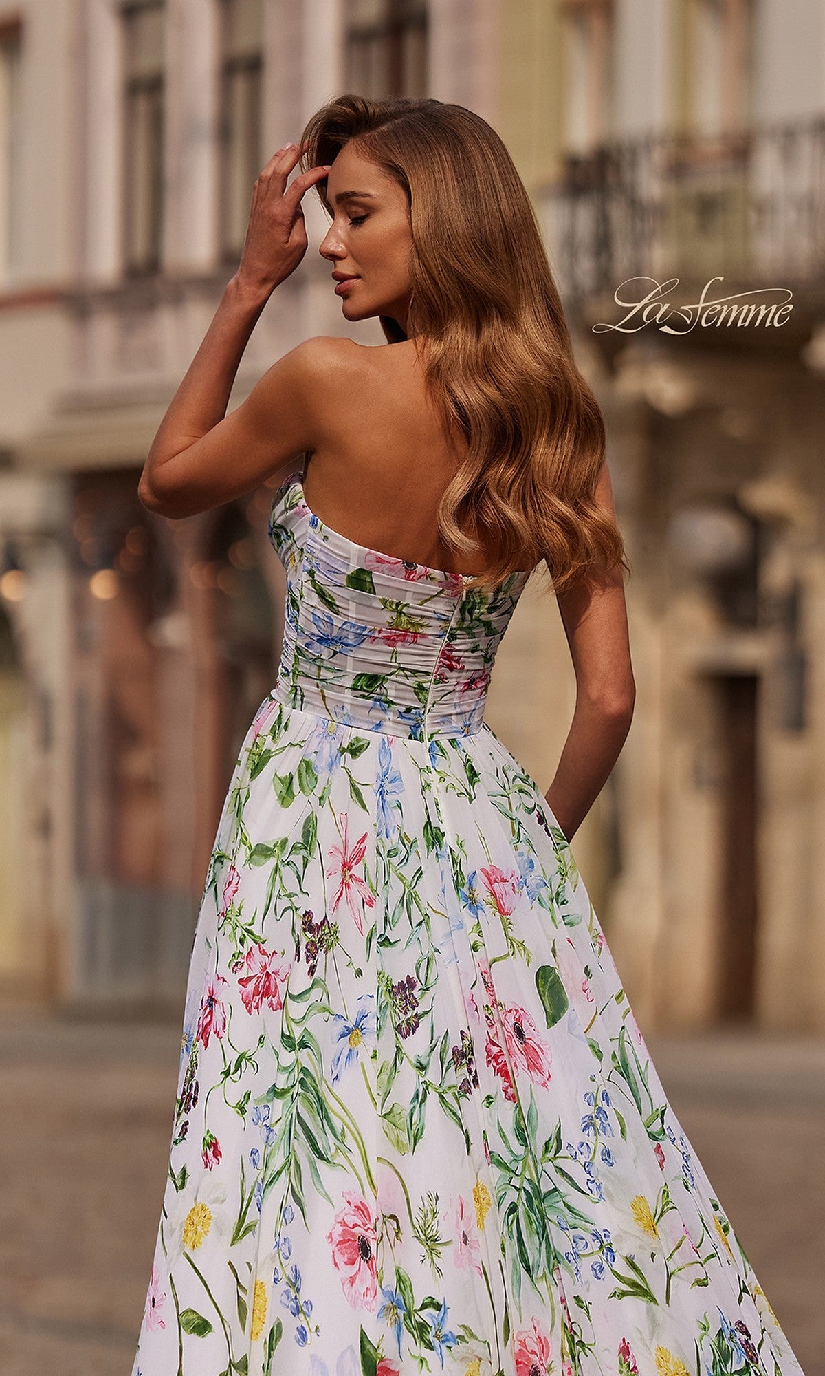 A woman with long wavy hair wears the La Femme 33450 long prom dress with a strapless floral a-line skirt, standing outdoors and facing away from the camera. 