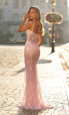 A woman wearing the Beaded-Lace Long Prom Dress, La Femme 33485—a fitted, floor-length pink gown with floral lace and thin straps—stands on a sunlit cobblestone street, looking back over her shoulder.