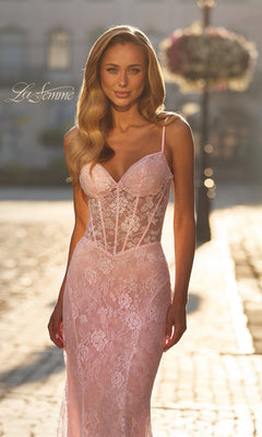 A woman stands outdoors on a sunlit cobblestone street in the La Femme 33485 Beaded-Lace Long Prom Dress, featuring delicate floral lace, thin straps, and a fitted sheer bodice with visible boning details.