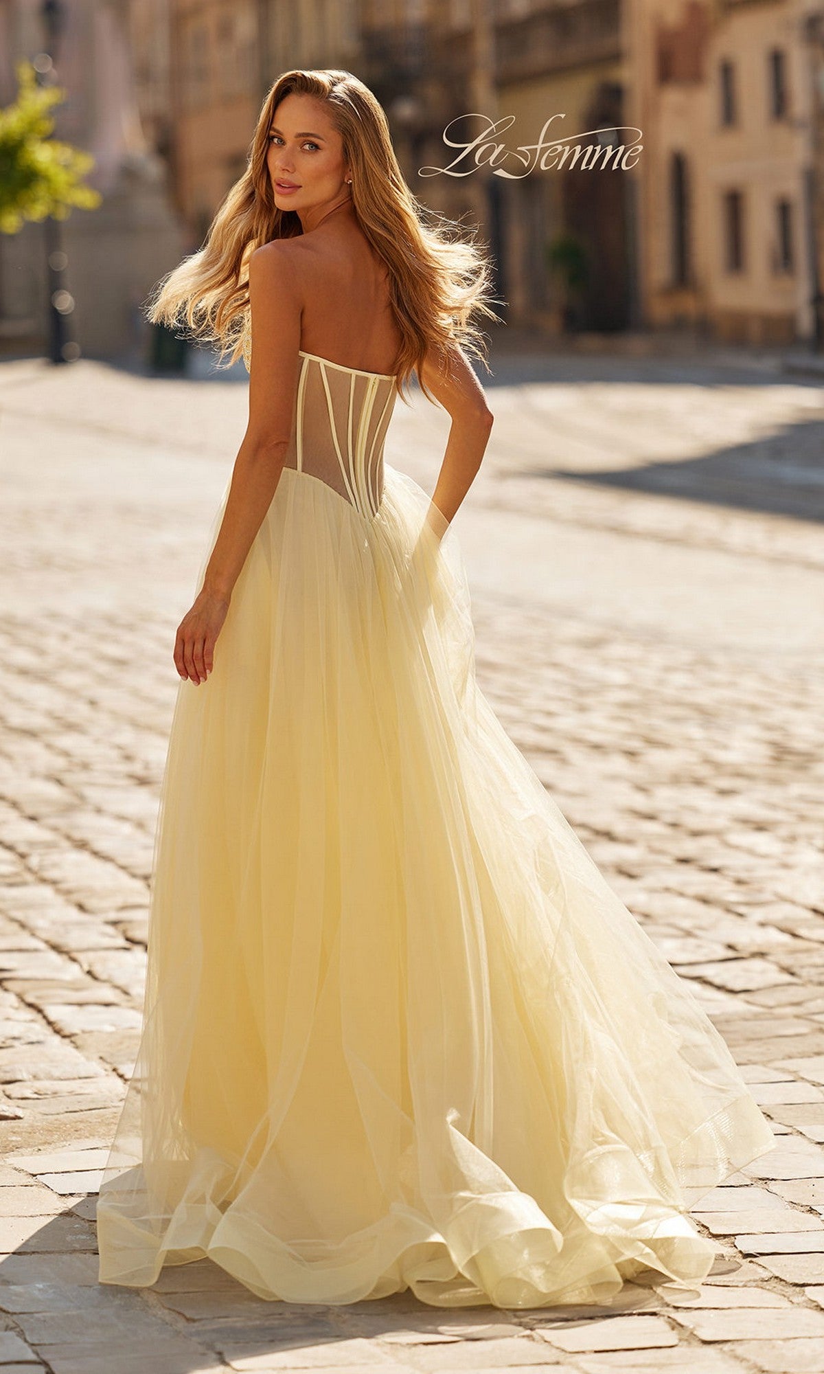 A woman walks on a sunlit cobblestone street wearing the La Femme 33491, a strapless sheer-corset prom gown with an embroidered bodice and long a-line skirt.