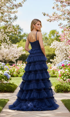 A woman stands on a garden path surrounded by flowers, wearing the La Femme 33493 long prom dress—a navy blue gown with spaghetti straps and a tiered ruffle skirt.